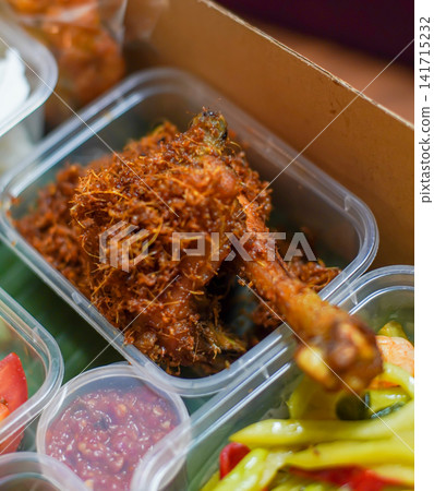 Crispy Indonesian fried chicken covered with fragrant fried galangal, served with spicy sambal and colorful pickled vegetables in takeaway containers, representing Southeast Asian cuisine. Crispy Indonesian fried chicken covered with fragrant fried galangal, served with spicy sambal and colorful pickled vegetables in takeaway containers, representing Southeast Asian cuisine. 141715232