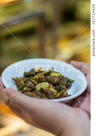 Authentic spicy stir fried chicken hearts with chili served in a bowl, Indonesian street food scene with hand holding and cooking tray background, rich color and texture 141715424