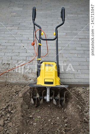 A close-up view of a yellow and black electric garden tiller actively working at the edge of a paved area and soil. The rotating metal tines dig into the earth, loosening and turning it for planting 141715847