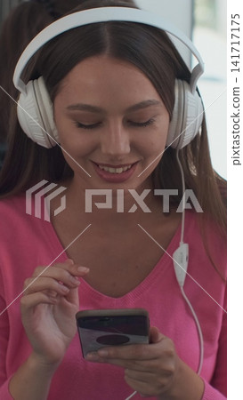 Vertical video. Young woman passenger enjoying trip at the public transport, sitting with headphones near the window in the modern tram. Vertical video. Young woman passenger enjoying trip at the public transport, sitting with headphones near the window in the modern tram. 141717175