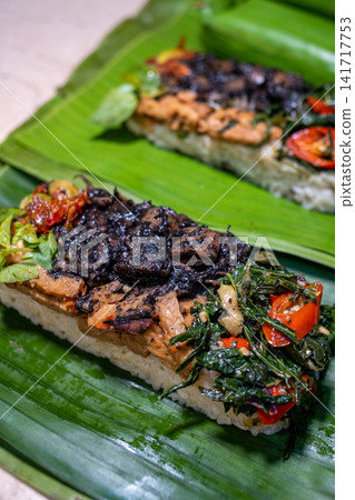 Traditional Indonesian seafood rice with black squid ink, vegetables, herbs, and sambal served on banana leaf. Authentic Southeast Asian cuisine suitable for travel, restaurant, and food publications. Traditional Indonesian seafood rice with black squid ink, vegetables, herbs, and sambal served on banana leaf. Authentic Southeast Asian cuisine suitable for travel, restaurant, and food publications. 141717753