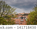 Scenic Prague cityscape framed by fresh spring trees with historic red rooftops and castle architecture beneath dramatic cloudy skies, capturing the timeless beauty of Prague City Czech Republic 141718430