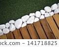 Detail of a garden pathway featuring artificial grass, wooden decking tiles and white pebbles along the border, creating a modern outdoor landscaping design. 141718825