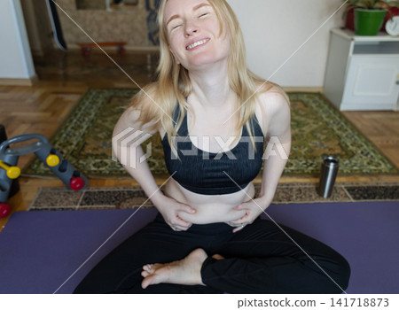 Smiling woman sitting on a yoga mat preparing for breathing exercises and weight loss practice, embracing her body positivity and postpartum belly at home. Smiling woman sitting on a yoga mat preparing for breathing exercises and weight loss practice, embracing her body positivity and postpartum belly at home. 141718873