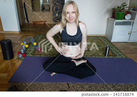 Smiling woman sitting on a yoga mat preparing for breathing exercises and weight loss practice, embracing her body positivity and postpartum belly at home. Smiling woman sitting on a yoga mat preparing for breathing exercises and weight loss practice, embracing her body positivity and postpartum belly at home. 141718874