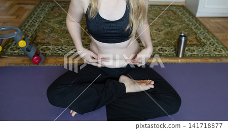 Smiling woman sitting on a yoga mat preparing for breathing exercises and weight loss practice, embracing her body positivity and postpartum belly at home. Smiling woman sitting on a yoga mat preparing for breathing exercises and weight loss practice, embracing her body positivity and postpartum belly at home. 141718877