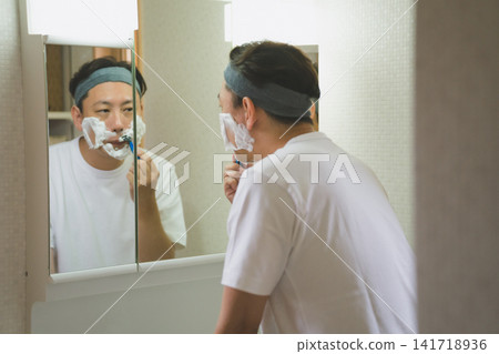 A man in his 40s shaves his beard while checking his reflection in the bathroom mirror. A man in his 40s shaves his beard while checking his reflection in the bathroom mirror. 141718936