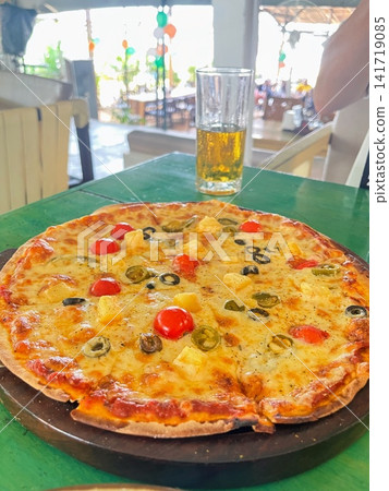 A large plate of pizza topped with cherry tomatoes and pineapple, served on a wooden board, along with a glass of drink. A large plate of pizza topped with cherry tomatoes and pineapple, served on a wooden board, along with a glass of drink. 141719085