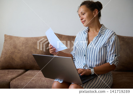 Woman working at home on laptop while reviewing a document on the couch Woman working at home on laptop while reviewing a document on the couch 141719289