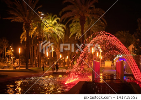 Nighttime park scene featuring illuminated palm trees and a colorful fountain with vibrant water jets in a public space Nighttime park scene featuring illuminated palm trees and a colorful fountain with vibrant water jets in a public space 141719552
