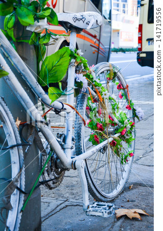 White bicycle adorned with green leaves and pink flowers parked against a pole on a city street, with blurred vehicles in the background White bicycle adorned with green leaves and pink flowers parked against a pole on a city street, with blurred vehicles in the background 141719556