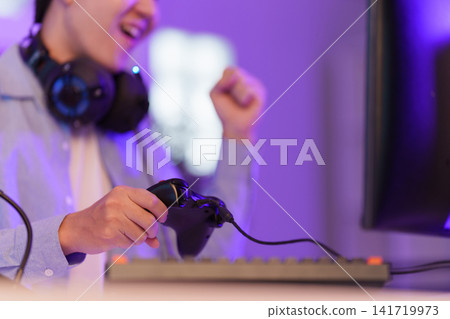 Asian gamer holding controller celebrating video game victory in purple room 141719973