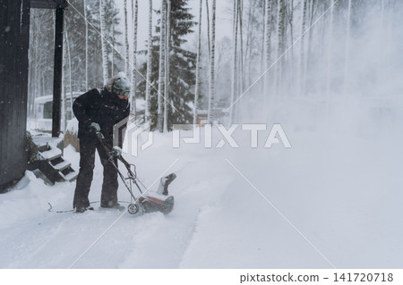 man clearing snow near wooden house in countryside using electric snow blower 141720718