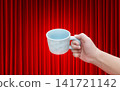An empty white ceramic coffee cup is in a man's hand, with a red curtain as a background. 141721142