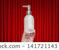 A clear glass perfume bottle with a diamond pattern is held in hand, against a red curtain as a backdrop. 141721143