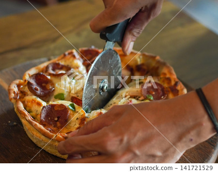 Freshly baked pizza with melted cheese being sliced by hand. Warm lighting and crispy crust create appetizing food image for restaurant and delivery concepts. Freshly baked pizza with melted cheese being sliced by hand. Warm lighting and crispy crust create appetizing food image for restaurant and delivery concepts. 141721529