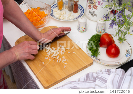 A person is peeling garlic cloves while preparing a salad in a kitchen. Fresh vegetables, spices, and utensils are arranged on a table nearby A person is peeling garlic cloves while preparing a salad in a kitchen. Fresh vegetables, spices, and utensils are arranged on a table nearby 141722046