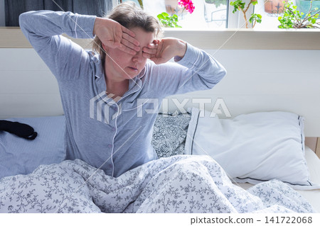 A 50-year-old woman appears tired and distressed as she sits up in bed. She rubs her eyes, indicating a lack of sleep, while surrounded by soft bedding in a bright room A 50-year-old woman appears tired and distressed as she sits up in bed. She rubs her eyes, indicating a lack of sleep, while surrounded by soft bedding in a bright room 141722068