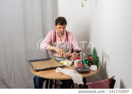 A 53-year-old man is preparing chicken for pre-Christmas dinner. He seasons the chicken in a bowl. Nearby are potatoes and a glass of wine on the table A 53-year-old man is preparing chicken for pre-Christmas dinner. He seasons the chicken in a bowl. Nearby are potatoes and a glass of wine on the table 141722097