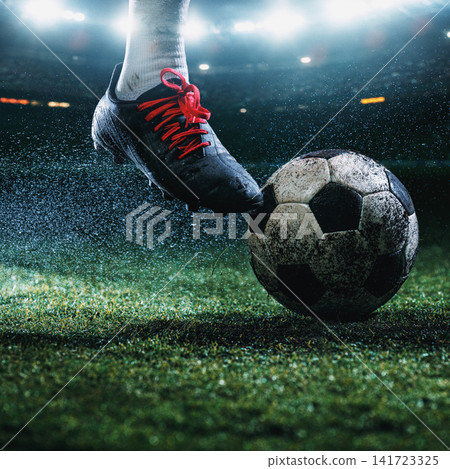 Red-laced football boot stopping dirty soccer ball on rain-soaked turf under bright stadium lights Red-laced football boot stopping dirty soccer ball on rain-soaked turf under bright stadium lights 141723325