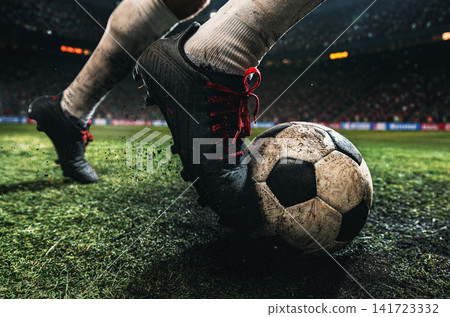 Low-angle close-up of muddy football boots controlling worn ball on textured stadium grass Low-angle close-up of muddy football boots controlling worn ball on textured stadium grass 141723332