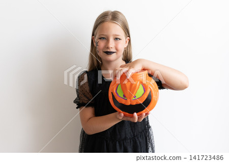 Girl in witch costume resting hand on pumpkin bucket on white background 141723486
