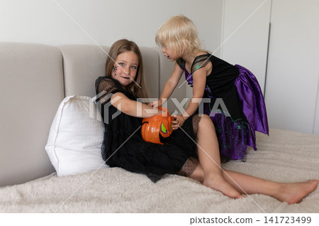 Sisters in witch costumes playing with pumpkin bucket on bed Sisters in witch costumes playing with pumpkin bucket on bed 141723499