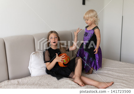 Girls in witch costumes gesturing surprised on bed with pumpkin bucket Girls in witch costumes gesturing surprised on bed with pumpkin bucket 141723500