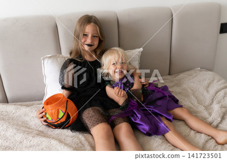 Two sisters in witch costumes hugging and laughing on bed with pumpkin bucket Two sisters in witch costumes hugging and laughing on bed with pumpkin bucket 141723501