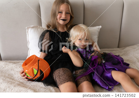 Sisters in witch costumes cuddling on bed holding pumpkin bucket Sisters in witch costumes cuddling on bed holding pumpkin bucket 141723502