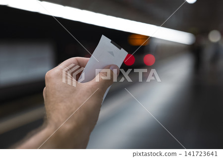 Hand holding a white ticket at a subway station platform, illustrating daily commuter interaction with modern transport systems. Hand holding a white ticket at a subway station platform, illustrating daily commuter interaction with modern transport systems. 141723641
