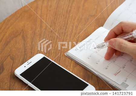 A woman's hand writing in a November schedule book and holding a smartphone on a wooden table. A woman's hand writing in a November schedule book and holding a smartphone on a wooden table. 141723791