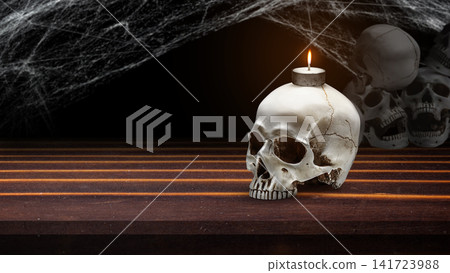 A spooky Halloween scene featuring a human skull candle holder with a burning tea light on a wooden table, decorated with cobwebs and additional skulls in the dark background. Concept of Halloween A spooky Halloween scene featuring a human skull candle holder with a burning tea light on a wooden table, decorated with cobwebs and additional skulls in the dark background. Concept of Halloween 141723988