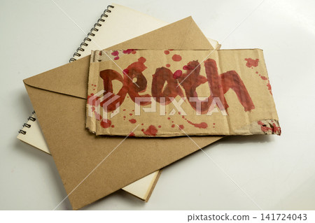 A piece of cardboard with the word death written in red blood, placed on top of a brown paper envelope and a spiral notebook, creating a dark, suspenseful concept. Concept of Halloween A piece of cardboard with the word death written in red blood, placed on top of a brown paper envelope and a spiral notebook, creating a dark, suspenseful concept. Concept of Halloween 141724043