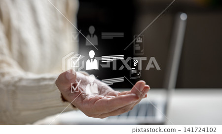 Female hand holding virtual HR icons representing recruitment, candidate selection, and user profiles with a laptop in the background. HR Recruitment concept 141724102