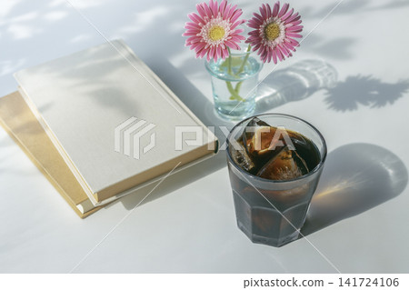 夏日閱讀的畫面:冰咖啡、粉紅色的非洲菊和一本白色精裝書。 夏日閱讀的畫面:冰咖啡、粉紅色的非洲菊和一本白色精裝書。 141724106