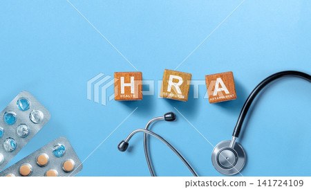 Wooden blocks spelling HRA for Health Reimbursement Arrangement alongside a stethoscope and pills on a blue background. Concept of Reimburse 141724109