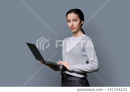 Young female student holding laptop computer and looking at camera on grey background Young female student holding laptop computer and looking at camera on grey background 141724223