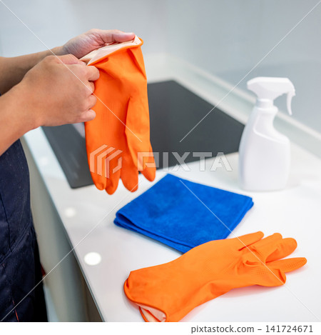 Hand putting on orange rubber gloves with blue microfiber cloth, and cleaner spray bottle nearby Hand putting on orange rubber gloves with blue microfiber cloth, and cleaner spray bottle nearby 141724671