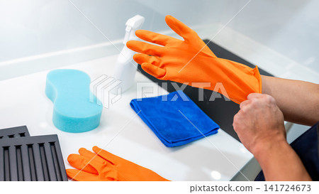 Hand putting on orange rubber gloves with blue microfiber cloth, sponge and spray bottle nearby Hand putting on orange rubber gloves with blue microfiber cloth, sponge and spray bottle nearby 141724673