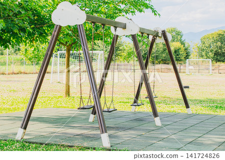 Empty wooden swing set with chain seats standing on soft rubber safety tiles at outdoor public kids playground with green trees and lawn background on sunny summer day Empty wooden swing set with chain seats standing on soft rubber safety tiles at outdoor public kids playground with green trees and lawn background on sunny summer day 141724826