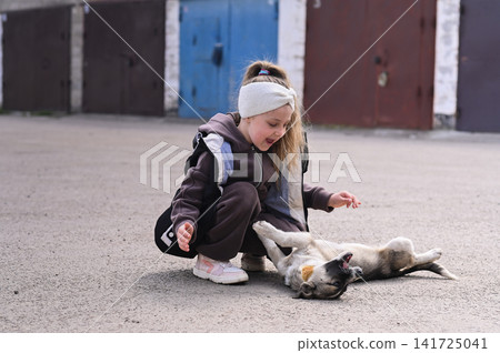 Little girl plays with stray dog on asphalt road near garages. Little girl plays with stray dog on asphalt road near garages. 141725041
