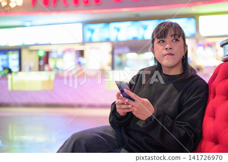 Asian Woman Pausing with Smartphone on Plush Armchair Asian Woman Pausing with Smartphone on Plush Armchair 141726970