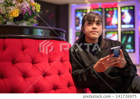 Asian Person Seated on Plush Couch Holding Smartphone Asian Person Seated on Plush Couch Holding Smartphone 141726975