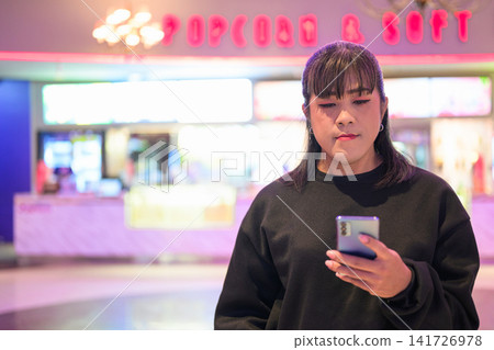 Asian Person Checking Smartphone near Cinema Concession Stand Asian Person Checking Smartphone near Cinema Concession Stand 141726978