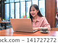 Smiling Asian Woman Waving at Her Laptop Screen during a Video Call 141727272