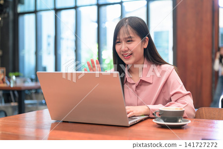 Smiling Asian Woman Waving at Her Laptop Screen during a Video Call Smiling Asian Woman Waving at Her Laptop Screen during a Video Call 141727272