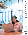 Asian Woman Having a Phone Call while Working on Her Laptop 141727273