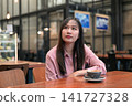 Thoughtful Asian Woman Looking Up while Sitting in a Cafe with Coffee 141727328