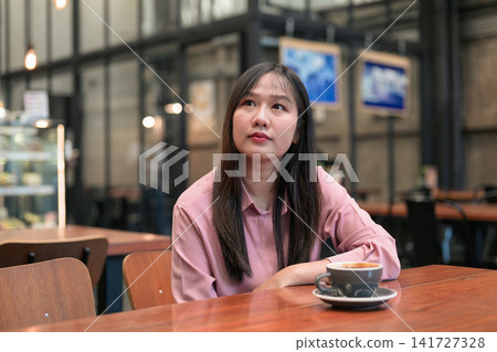 Thoughtful Asian Woman Looking Up while Sitting in a Cafe with Coffee Thoughtful Asian Woman Looking Up while Sitting in a Cafe with Coffee 141727328